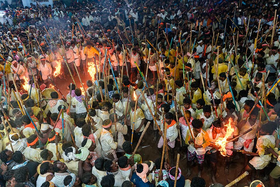A dense crowd of devotees holding long bamboo sticks and torches at night during the Banni festival